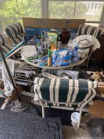 Photo shows round outdoor table with metal frame and glass center, cluttered with various items. Four outdoor metal chairs around table with green and white striped cloth covers on seats and backs.
