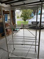 White collapsible metal shelving unit with four wire wire grid shelves. It features a tall frame with wire grid shelves placed evenly vertically. The shelving is standing in a garage with natural daylight.