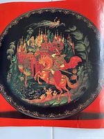 Close-up image of one decorative plate showing detailed hand-painted scene of Ruslan on horseback with Russian architecture and ornamental foliage in black and gold border.