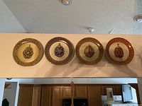 Photo of four framed Franklin Mint collector plates featuring jeweled eggs hanging on a wall above kitchen cabinets. Plates have ornate decorative borders and detailed egg designs in different color schemes.