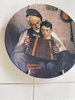 Close-up photo of one Norman Rockwell plate featuring an elderly man teaching a young boy to play an accordion.