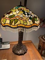 The photo shows a Tiffany style table lamp featuring a grapevine patterned stained glass shade in various colors including green, white, purple, and amber. The shade has a clear bead fringe along the bottom edge. The lamp base is ornate and bronze-toned, resting on a wooden table.
