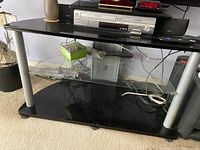 TV stand with black metal and plastic structure, middle clear glass shelf, cables and electronic devices on top and beneath.