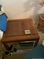 Square wooden side table with rounded edges and medium brown finish shown from top and side angle. A small coaster sits on top. Table is in carpeted room next to other furniture.