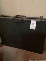Closed black rectangular portfolio case with handle and metal clasps, propped against a wall.