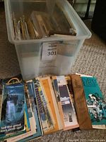 Photo showing a clear plastic bin filled with numerous old maps and brochures standing up vertically inside. Several colorful travel brochures are fanned out next to the bin, visible with images and text on the covers.