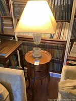 Round wooden end table under lit glass table lamp, showing the top and general shape of the table next to bookshelves.