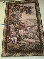 Full view of the tapestry hung on the wall showing the fox hunt scene, riders with dogs and a tree on the left, with hills in the background.