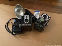 Photo showing the Yashica 635 camera with twin lenses and the Kodak Brownie Flash camera with attached flash bulb reflector and black handle, placed on wooden floor.