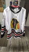 Front view of white Chicago Blackhawks jersey showing embroidered Native American head logo and black/red striped sleeves.