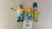 Front view of three bendable Simpsons figures - Homer, Bart, and Marge, placed upright on a surface with lot number sign.