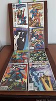 Photo showing eight Superman comic books arranged on a wooden table, featuring varied cover art including Superman with a child, Superman with other characters, dark shadowed face art, and action scenes.