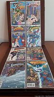 Top-down view of eight Superman comic books arranged in two rows on a wooden floor, showing varied colorful illustrated covers including scenes of Superman and other characters.