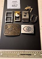 Top front view showing seven items: six metal buckles and a miniature pistol item, displayed on a black surface with a ruler for scale.