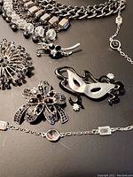 Close-up of jewellery pieces showing a masquerade mask brooch, floral brooches, and chains with rhinestones on black background