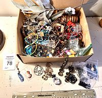 Box with assorted jewelry parts and craft items including beads, chains, pendants, and decorative pieces spread inside and outside the box, with ruler for scale.