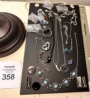 Top view photo showing assorted jewelry items including necklaces with pendants, brooches, earrings, and small plastic bags with jewelry parts on a black display board with a ruler for scale.