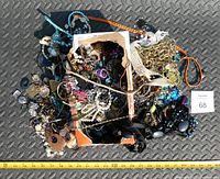 Top-down image showing assortment of craft and costume jewelry in a box and spread around it on a textured dark surface with measuring tape for scale.