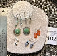 Top view of six pairs of earrings arranged on a textured white surface with a ruler for scale, showcasing various styles and semi-precious stones including turquoise and coral.