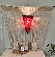 Overall view of the red beaded hanging light with string fringe bundles beneath it; light is turned on showing glow and shadows