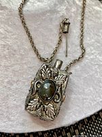 Close-up of a silver-tone chain necklace with a rectangular pendant featuring embossed leaf designs and a central oval green stone. Includes a pin accessory beside the chain.