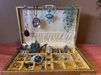 View of jewelry lot in storage box showing necklaces hanging on lid and rings, watch, bracelets, and small jewelry items in compartments