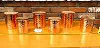 Front view of all five copper-toned hammered metal canisters with wooden lids, arranged in a row from smallest to largest on a wooden surface with a brick background.