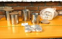 Four silver-toned hammered metal canisters of varying sizes with flat wooden lids, plus a set of six small silver-toned containers with lids, displayed on a wooden surface against a brick background.