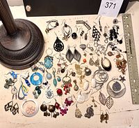 Photo of a large variety of single earrings displayed on a white surface with a ruler showing scale and part of a lamp base visible.