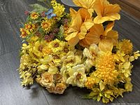Cluster of mixed yellow, orange, cream artificial flowers with some green foliage on dark flooring.