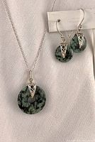 Close-up of necklace pendant and matching earrings against white fabric background showing green and black speckled semi-precious stone with silver swirl accents