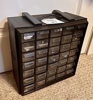 Black plastic parts organizer with 30 transparent drawers arranged in five rows of six drawers each on a carpeted floor.