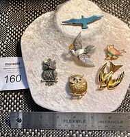 Six bird-themed pins and brooches displayed on a white holder with a flexible ruler showing scale. Includes blue enamel bird, silver dove, gold owls, and swallows with enamel flowers.