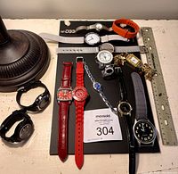 Lot displayed on black board with ruler for scale including diverse 10 wristwatches and a decorative gold-tone bracelet watch shown with charms.