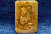 Front side of rectangular light brown jade pendant with carved image of a woman holding a bird, framed by architectural background elements.
