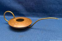 Side angle showing the long spout, wide circular copper body, and brass handle of the watering can on a blue fabric background.