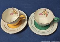 Front view of two Aynsley demitasse teacups and saucers on blue background, showing floral interior and gold trim