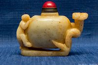 Side view of the small pale yellowish-green carved jade opium snuff bottle showing a reclining carved human figure on the body and the red rounded stopper on top.