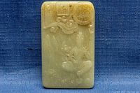 Front view of the rectangular jade pendant showing the carved embossed figure and cloud motifs on blue fabric background.