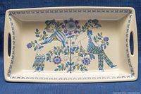 Top angled view of rectangular ceramic serving dish with colorful stylized figures and floral motifs in blue and green, handles on short sides visible.