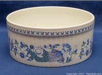 Side view of round ribbed ceramic baking dish showing blue floral and figure design by Lotte Turi for Figgjo, on a blue fabric background.