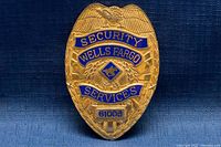 Front view of gold-colored Wells Fargo Security Services metal badge with embossed shield shape and blue lettering.