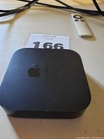 Top view of black Apple TV box with Apple logo and 'tv' lettering. Accompanied by white remote and cords on a light surface.