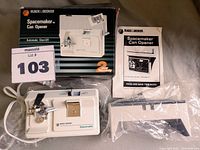 Full view of Black & Decker Spacemaker can opener unit, instruction manual, and plastic mounting bracket on table with original box showing some damage.