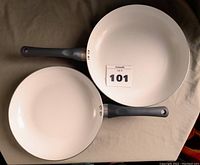 Two ceramic non-stick frying pans placed side-by-side on a surface, showing white interiors and black handles.