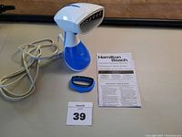 Full view of Hamilton Beach handheld clothes steamer, small brush attachment, and instruction manual with power cord coiled behind steamer