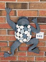 Full view of metal frog wall décor mounted against brick wall, showing entire frog shape and decorative body.