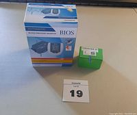 Photo shows BIOS Automatic Blood Pressure Monitor box and a small green unopened box containing magnets, placed on a light surface with lot number card 19.