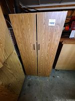 Closed view of two door wooden pantry cabinet with wood grain finish and black handles.