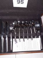 Close up of silverware pieces arranged inside the velvet-lined wooden case, including various knives, forks, and spoons.